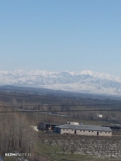 A wide landscape features a clear blue sky above snow-capped mountains in the distance. A flat terrain stretches towards the mountains, marked by rows of bare trees and fields with scattered patches of snow. In the foreground, a long, low building with a gray roof stands amidst a bare orchard, its doors and windows visible. Power lines cross the scene horizontally. The text "REDMI NOTE 12" appears at the lower-left corner.