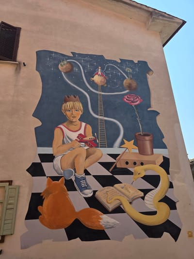 This is a mural painted on the side of a building in Viterbo, Italy, depicting a scene inspired by "The Little Prince." In the foreground, a young boy with blond hair and a crown sits on a black and white checkered floor, holding a red toy airplane. To his left is an orange fox with its back to the viewer. To his right, a yellow snake is coiled around an open book, its tongue flicking out. The book contains handwritten text and an illustration. Nearby are a wooden box with holes, a golden star wand, and a potted rose. In the background, the sky is dark blue, filled with stars. Several planets are depicted, with small figures on them, including one on a planet connected by a ladder, reminiscent of the aviator's encounter with the Little Prince. A single red rose in a pot is also visible. The mural is on a stucco wall, with a portion of a window with wooden shutters visible to the left of the mural. The sky above is clear and blue.