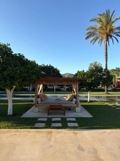 The image captures a serene outdoor scene, possibly a resort or a luxurious garden in Santa Eulària des Riu, Spain. The primary subjects are a wooden gazebo with comfortable seating, a neatly manicured lawn, and a swimming pool area.
The gazebo, situated on a concrete base with stepping stones leading to it, features light-colored cushions and pillows, suggesting a place for relaxation. Behind the gazebo, lounge chairs are visible by the pool, hinting at leisure activities. A tall palm tree dominates the left side of the frame, reaching towards a clear blue sky, indicating fair weather and likely daytime, possibly late afternoon given the warm lighting. The presence of lush green trees and what appears to be a distant mountain range in the background further defines the natural setting.
The overall mood is tranquil and inviting, emphasizing a peaceful escape. The concrete pathway on the right side of the image, with its textured surface and slight discoloration, suggests an outdoor living space or patio. No people are visible in the frame, contributing to the sense of quietude. There is no visible text in the image.
