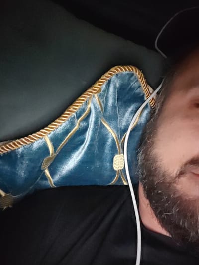 A man with a beard, wearing a black t-shirt, is lying down with a plush blue pillow with gold trim under his head. A white cord, possibly from headphones, rests on his ear and cheek. The scene is dimly lit, suggesting an indoor setting, possibly a bedroom. The man appears relaxed. The location is not discernible from the image.
