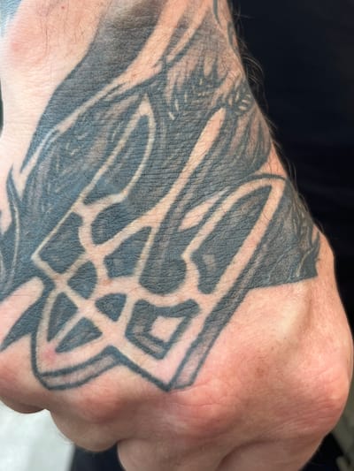 The image shows a close-up of a person's hand with a black tattoo on it. The tattoo appears to be a stylized trident, which is the national symbol of Ukraine. The tattoo covers a significant portion of the back of the hand and extends up onto the wrist. The skin is fair and shows some texture. The background is dark and out of focus. The image does not contain any information about the location in the city of Palanca, Moldova.