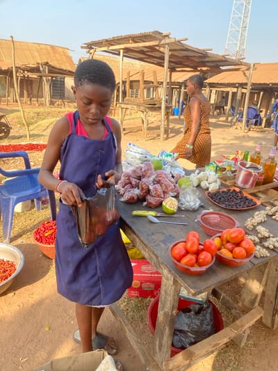 A young woman wearing a purple apron stands at a wooden table laden with produce, packing items in a plastic bag. The table displays tomatoes, ginger, limes, plums, and bagged items, likely peppers or spices. In the background, another woman stands nearby. Stalls with corrugated iron roofs suggest an outdoor market setting. Pails of red peppers sit on the ground, adding to the market atmosphere. A communications tower is visible in the distance.