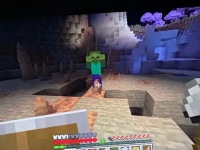 This is a screenshot from the video game Minecraft, depicting an in-game character, a zombie, standing in a cave-like environment. The zombie is green with purple pants and a teal shirt. The environment features stone blocks, some with orange moss, and purple, mushroom-like flora in the background. The player's user interface is visible at the bottom of the screen, showing health bars, a hunger bar, and inventory items. The location is not a real-world city like Bezden, Bulgaria, but rather a virtual environment within the game.