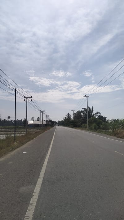 The image depicts a straight asphalt road stretching into the distance under a partly cloudy sky. The road is bordered on the left by a grassy shoulder and a rice paddy. On the right, the road is lined with vegetation, including palm trees and tall grass. Utility poles with power lines run along both sides of the road, suggesting a rural or semi-rural setting. In the background, a few buildings are visible, and a vehicle can be seen in the distance on the road. The scene appears calm and typical of a roadside in a tropical region. The location is identified as Kruengseupeng, Indonesia.