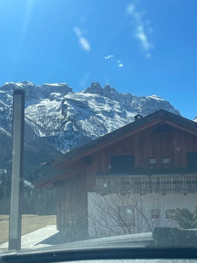 A landscape photograph depicts a snow-dusted mountain range against a bright blue sky with sparse clouds. The image is taken from within an interior space, indicated by the presence of building structures including a chalet-style building on the right and a metal window frame along the bottom edge. The perspective is tilted. The location is suggested as being in Sappada, Italy based on the prompt. There is no visible human activity, people, subjects, or interactions.