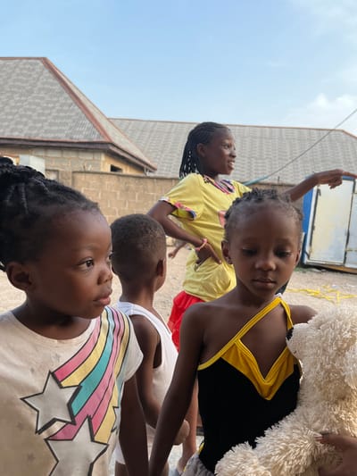 A group of four children are standing outdoors in Iddah, Nigeria. The background includes buildings constructed of gray blocks and a clear, light blue sky. The child in the foreground is wearing a white shirt with a colorful rainbow and star design. Behind this child is another wearing a plain white tank top. A third child is wearing a yellow shirt and has their arm raised, while the fourth child is in a black and yellow swimsuit and holding a white, fluffy stuffed animal.
