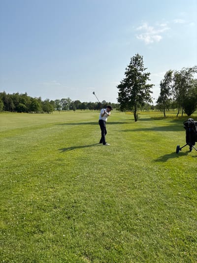 The image shows a golfer in a white shirt and black pants swinging a golf club on a golf course. The golfer is facing right and has a black cap on their head. They are standing on a green, grassy fairway. There is a black golf bag on wheels in the background, and a large tree to the right of the frame. There are trees and bushes lining the fairway in the distance. The sky is blue and clear. The golfer is located at Pocklington Golf Club in Pocklington, UK. The location can be identified using the latitude and longitude coordinates provided: 53.939, -0.76.