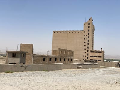 The image shows a partially constructed building complex in Panjwai, Afghanistan, at coordinates 31.614 latitude and 65.588 longitude. The complex is made of concrete blocks and consists of two buildings. The larger building is a tall, rectangular structure with multiple levels, while the smaller building is a single-story structure with several rooms. The smaller building is located in the foreground, while the larger building is in the background. The larger building has a section of the roof missing. There is a concrete wall in front of the buildings, with a sign written in an unknown language. The ground is covered in gravel. The sky is clear and blue. No people or other objects are visible in the image.