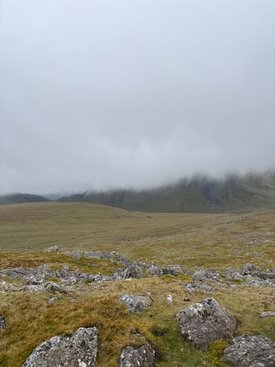 The image shows a mountain landscape in Llanberis, United Kingdom. The right side of the image features a steep, rocky slope covered in grass and moss. Large grey rocks are scattered across the slope. To the left, a grassy mountain descends into a dense layer of fog. A faint trail or path is visible on the grassy hillside, suggesting hikers may be present, although no individuals are clearly visible. The overall atmosphere is misty and overcast.