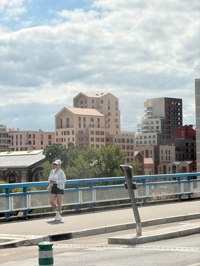 A young woman, wearing a white cap, a light blue jacket, and black shorts, stands on a sidewalk next to a blue metal fence. She is looking down at something in her hands. The background features a series of modern buildings with geometric designs and various shades of beige and red. Some buildings have balconies and many windows. Green trees are visible between the buildings. The sky is overcast with white clouds. A lamppost is attached to the fence. The sidewalk is paved with light gray concrete and bordered by a darker gray curb. The time of day appears to be daytime. The overall impression is that of an urban environment on a cloudy day.