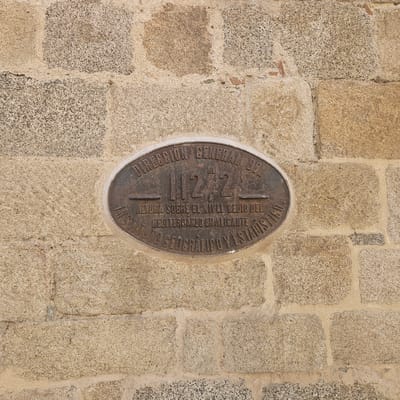 The image shows an oval, bronze-colored plaque affixed to a rough stone wall, likely part of a building. The plaque bears the inscription "Dirección General del 112,2 m. Altura Sobre el Nivel Medio del Mediterráneo en Alicante, Instituto Geográfico y Estadístico". The inscription is in Spanish. The wall is constructed of irregular, light-colored stone blocks joined with mortar. The overall scene suggests an official marker indicating the altitude above sea level, placed by the Spanish Geographical and Statistical Institute in reference to the average Mediterranean sea level in Alicante. The location is in the city of La Jonquera, Spain.
