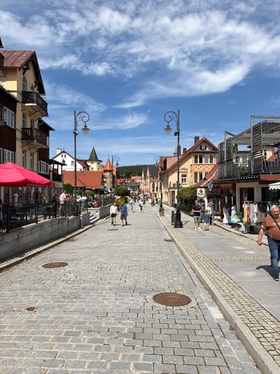 The image depicts a street scene in Świeradów-Zdrój, Poland, characterized by a cobblestone street lined with buildings on both sides. The architecture appears to be traditional with elements like wooden balconies and pitched roofs, some painted in yellow and red. Several people are walking on the street and sidewalks. On the left, there is a building with a pink umbrella in front of a shop. On the right, businesses display clothing racks outside their establishments. Street lamps with ornate designs are positioned along the street. The sky is blue with scattered clouds, suggesting fair weather. The overall atmosphere is that of a bustling pedestrian area in a town.