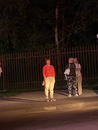 The media file shows two women standing outdoors at night, with a fence behind them and a sidewalk in front. The woman closer to the fence is wearing a white t-shirt with text that appears to say "WE ALL GOOD". She is also wearing light-colored pants. The woman in front of her is wearing an orange long-sleeved shirt and light-colored pants. Both women appear to be middle-aged. A white mask is hanging from the fence near the woman in the white t-shirt. The scene is dimly lit, suggesting it is nighttime. There are trees visible to the left of the fence, and the ground in front of the women appears to be a sidewalk or paved area.