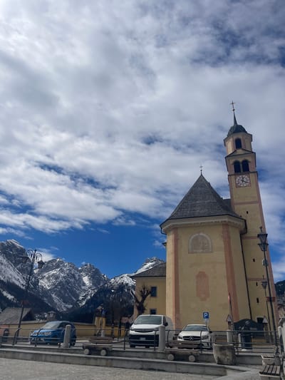 The image shows a church in Sappada, Italy, with a tall tower and clock. The church is painted a light yellow with red trim. There is a mountain range visible in the background, with snowy peaks. Several parked cars are in front of the church. The sky is blue with white clouds.