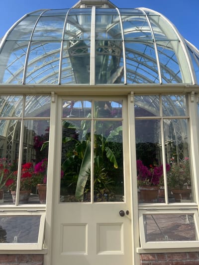The image shows the exterior of a large, ornate greenhouse, likely part of Ardgillan Castle in Ireland. The greenhouse has a domed glass roof and glass walls, with a cream-colored frame. Through the glass, numerous potted plants and flowers are visible, including red and pink flowering plants, and large-leafed tropical plants. The structure sits on a brick base. The sky is a clear, bright blue. The scene appears to be taken on a sunny day.