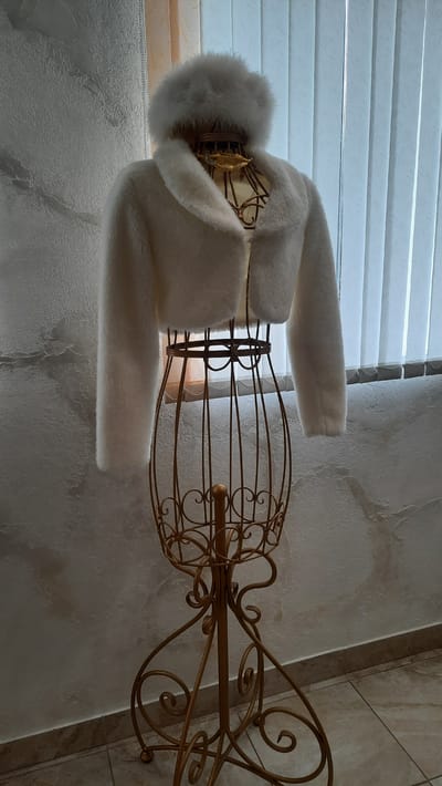 A white faux fur jacket and matching hat are displayed on a gold wire mannequin in what appears to be an indoor setting. The mannequin has an ornate, vintage design. Behind the mannequin, vertical blinds are visible, suggesting a window. The wall behind the mannequin has a textured, possibly plaster or stucco finish, with some discoloration. The floor is tiled. The overall impression is of a display for clothing or accessories. The provided information about the location in Belarus does not appear to be directly deducible from the visual evidence in the image.