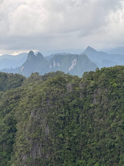 The image depicts a panoramic view of lush, green mountainous terrain, likely in the Khao Tang district of Thailand. The foreground is dominated by steep, densely forested slopes characterized by rugged, rocky outcrops interspersed with a thick canopy of trees. The vegetation appears verdant and untamed, suggesting a natural and somewhat wild environment. In the mid-ground and background, several distinct peaks rise, their shapes softened by atmospheric haze and distance. These mountains are also covered in trees, though the detail becomes less discernible with depth. The sky above is overcast, with muted, cloudy tones that contribute to a soft, diffused light across the landscape. The overall impression is one of a remote, natural landscape under a cloudy sky.