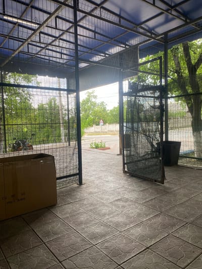 This image depicts an outdoor area with a caged structure, possibly a covered walkway or storage space, in Corjova, Moldova. The structure has a blue tarp roof supported by a metal frame and wire mesh walls. In the foreground, a cardboard box with text indicating "ITEM NO. H-994A" and "QTY: 108 PCS" sits on a tiled floor. The entrance of the caged area opens to a street scene with a white brick wall, trees, and a road. A traffic sign is visible in the distance. A dark-colored trash receptacle is placed near the right entrance of the caged area. The overall impression is of a functional, somewhat utilitarian outdoor space.