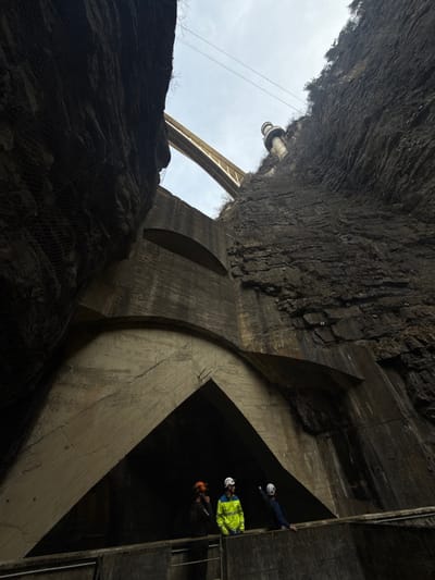 The image depicts a deep, narrow gorge in Pellafol, France, characterized by steep, dark rock walls and substantial concrete structures under an overcast sky.

In the lower and mid-ground, a massive concrete wall features multiple arched openings. The largest, central opening forms a dark, triangular entry point, flanked by a lighter, curved concrete arch above it. Another, smaller horizontal arched opening is visible higher up on the main concrete wall. The concrete exhibits signs of age, including stains and visible formwork patterns. On the left rock wall, a dark mesh net system is installed for rockfall protection.

Above the concrete structures and spanning the gorge, a light-colored arch bridge is visible against the grey sky. High on the right-hand rock face, a slender, cylindrical concrete tower with a rounded top protrudes upwards. Overhead, utility cables traverse the scene between the cliffs.

On a concrete walkway or platform in the foreground, three individuals are present. All are wearing hard hats: one in orange, two in white. The leftmost person wears dark clothing. The central individual wears a bright yellow-green high-visibility jacket and dark pants. The rightmost person wears a blue jacket and dark pants, and is captured in the act of pointing with their right arm towards the right rock wall, while looking in that direction. The overall lighting is dim due to the depth of the gorge and the overcast conditions.