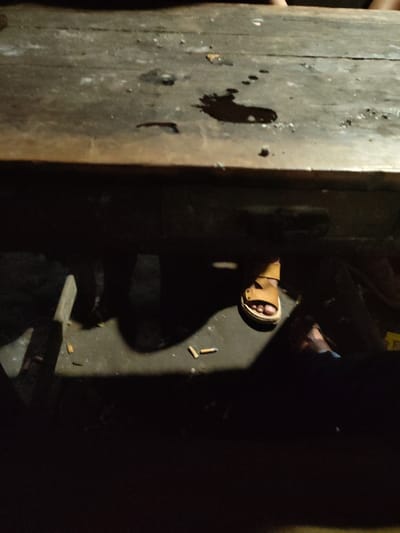 This image captures a low-light scene beneath and around a wooden table in Balla, Bangladesh. The upper portion of the frame displays a worn, dark wooden tabletop with multiple stains, including a prominent dark liquid spill resembling dried blood or oil towards the right, and numerous smaller splatters. Below the tabletop, the side of the wooden table structure is visible. The lower half of the image reveals a dark floor, likely concrete, illuminated by a localized light source from the upper left, casting strong shadows. On the floor, a person's bare foot, wearing a light brown open-toed sandal, is visible towards the right. Another darker, less distinct foot or leg is partially discernible further to the left, primarily within shadow. Several discarded cigarette butts are scattered on the floor directly beneath the table. No direct interactions or actions are discernible other than the presence of individuals' feet under the table. The environment appears utilitarian and somewhat disheveled.
