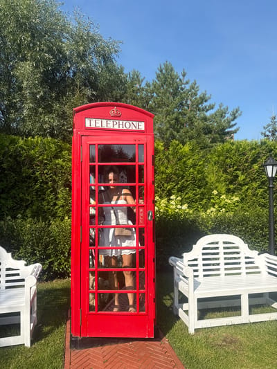 A woman in a white dress stands inside a red telephone booth in a garden setting. The telephone booth is a classic British design with "TELEPHONE" written across the top. The woman is reflected in the glass, and a beige purse is slung over her shoulder. Two white benches flank the telephone booth, one to the left and one to the right. Behind the booth and benches are manicured green hedges, tall trees, and a clear blue sky. A black lamppost stands to the right of the booth. The ground in front of the booth is paved with red bricks, and the rest of the area is covered in green grass. The image is taken in the city of Seltso, Russia, at a garden that appears to be a recreational area or a themed attraction.