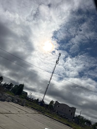 The image depicts an outdoor scene with a dramatic sky and an industrial structure. The primary subject is a tall, lattice transmission tower, angled diagonally across the frame, with several antennas attached to its upper section. Below the tower, a dilapidated building with a curved, damaged facade is visible. Construction materials, including stacks of concrete blocks, are scattered on the ground near the building. Trees and vegetation surround the structures, suggesting a semi-rural or industrial fringe location. Power lines are also prominent, stretching across the sky and connected to the tower.

The sky is filled with a mix of dark, ominous clouds and brighter, wispy formations. The sun is visible, peeking through the clouds on the left side of the image, creating a slight lens flare and a rainbow-like halo effect. This suggests it is daytime, likely late morning or early afternoon, with the weather being overcast but with moments of sunshine. The overall mood is somewhat somber and desolate, due to the decaying building and the heavy sky.

The image appears to have been taken from a slightly elevated or angled perspective, possibly from a vehicle. The ground in the foreground is paved with concrete slabs, showing wear and tear. No people are visible in the frame. There is no visible text in the image. The context provided indicates the location is Parma, Russia.