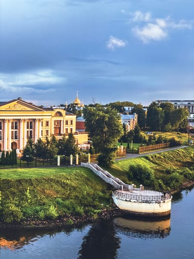 The image depicts a waterfront scene in the city of Novinki (Istrinsky District), Russia. In the foreground, a wide river occupies the lower portion of the frame, with its dark water reflecting the muted colors of the sky and surrounding environment. Emerging from the riverbank is a circular, elevated platform with a decorative railing. Two individuals are visible on this platform, one standing and looking out, the other seated or leaning. A curved staircase, also part of the waterfront infrastructure, ascends the grassy embankment from the platform.
The embankment itself is covered in lush green grass and dense foliage. Above the embankment, a long, stately building with a yellow facade and classical architectural elements, including columns and ornate pediments, is prominent. This building appears to be a significant structure, possibly a palace or administrative building. Behind it, other buildings with visible golden domes suggest a religious or historical complex. Further in the background, a tree-lined street runs parallel to the river, with a few vehicles visible, including a dark-colored car. The sky above is a mix of deep blue and heavy gray clouds, indicating potentially overcast or transitioning weather. The overall scene conveys a sense of historical grandeur and natural beauty.