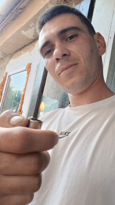 This media file captures a close-up, low-angle selfie of a man. He is wearing a white t-shirt with the Nike logo visible on the left side of his chest. The man is holding a small, metallic object, possibly a tool or component, in his left hand, which is positioned in the foreground and partially obscures his face. His right hand is not visible.

The background suggests an outdoor setting, possibly under construction or renovation. A window frame with expanding foam insulation is visible. The wall surrounding the window appears weathered and textured, with peeling plaster. The overall lighting indicates it is daytime. The man's expression is neutral, perhaps slightly curious or engaged with the camera. There are no clear indicators of specific weather conditions. The location context provided is Pishchanka, Ukraine.