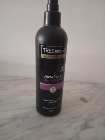 The image shows a black bottle of TRESemmé hair product with a spray top. The bottle is upright and positioned on a marble-like surface. Text on the bottle is visible in various colors, including gold, white, and purple. Some of the visible text includes "TRESemmé", "USED BY PROFESSIONALS", "Fortalece & REPARA", "CON FILTRO UV", "PARA UN CABELLO SUAVE Y BRILLANTE", and "PROTECTOR DE CALOR 300 ML". The background is a textured, light-colored wall. The image does not provide any information about the location in the city of Lalín, Spain, or any people, actions, or interactions.