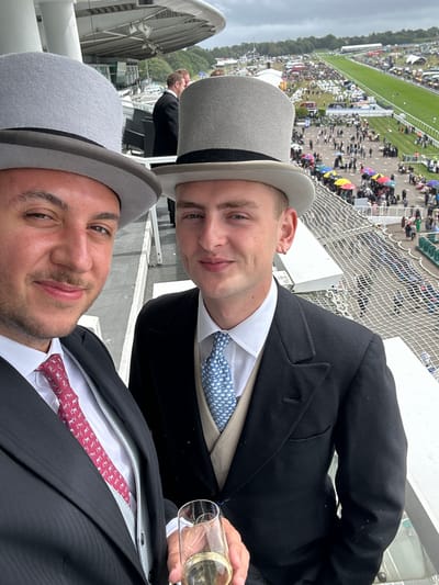 The image displays two men, positioned at an elevated vantage point, engaging in a selfie with a horse racing event unfolding in the background. The man in the foreground, on the left, possesses dark hair and facial stubble, wearing a grey top hat with a black band, a dark suit jacket, a white shirt, a grey waistcoat, and a red tie patterned with white horses. He holds a clear champagne flute containing a light yellow liquid in his right hand. The man on the right, slightly behind, has light hair and is clean-shaven, with a gold hoop earring visible in his left ear. He is attired in a grey top hat with a black band, a dark suit jacket, a white shirt, a beige waistcoat, and a light blue tie patterned with white ducks. Both individuals are looking towards the camera with neutral to slightly smiling expressions.

Behind the men, a vast outdoor scene unfolds, featuring a green horse racing track in the mid-distance. The track is surrounded by a large crowd of spectators, many gathered under colorful umbrellas in shades of red, yellow, and blue. Various temporary structures, including tents and marquees, are visible across the grounds. To the far left, a segment of a modern grandstand with a curved roof and white support columns is discernible. The sky above is overcast and grey. The overall environment, characterized by formal attire, a horse race, and grandstand architecture, places the scene at the Epsom Downs Racecourse in Epsom and Ewell, United Kingdom.