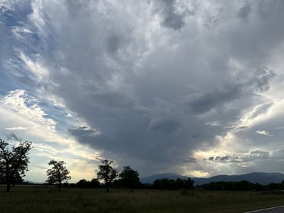 The image depicts a dramatic sky over a rural landscape, likely near Kran, Bulgaria, at dusk. The sky is dominated by a massive, dark, storm-like cloud formation in the center, with lighter, wispy clouds and patches of blue sky on the left and bright, sunlit clouds on the right. Rays of sunlight are breaking through the clouds on the right side, illuminating the distant mountain range and a line of trees. In the foreground, silhouetted trees stand in a grassy field. The overall impression is one of an approaching storm or the aftermath of one, with a striking contrast between dark and light. There are no people or specific identifiable landmarks that would pinpoint the exact location within Kran.