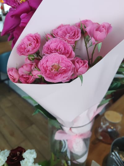 A close-up, high-angle shot captures a bouquet of vibrant pink roses and a large, deep pink peony. The flowers are elegantly wrapped in pale pink paper, with a delicate pink ribbon tied around the vase that holds the bouquet. The background is softly blurred, revealing a wooden floor and a glass jar containing some items. The scene appears to be indoors, possibly in a domestic setting or a flower shop, with no discernible connection to Amursk, Russia.