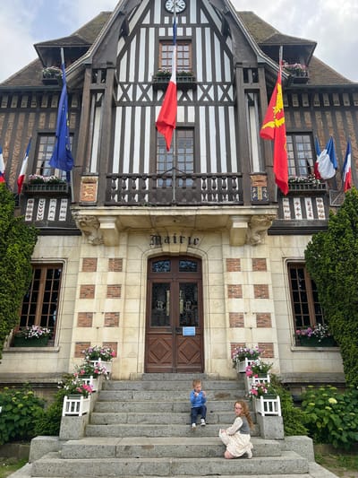 This is a full, eye-level shot of the exterior of the Hôtel de Ville, the town hall, in Deauville, France. The building is constructed in a traditional half-timbered Norman architectural style, with dark wooden beams against white plaster. The entrance is a large wooden door with ornate glass panels, flanked by stone and brickwork. Flags are displayed prominently on the facade, including the European Union flag, the French flag, and what appears to be the flag of Normandy.
In front of the town hall, a wide stone staircase leads up to the entrance. Two children are sitting on the stairs. A young boy in a blue sweater and dark pants is sitting on the fifth step from the bottom, looking down at something in his hands. A young girl with curly brown hair, wearing a patterned dress and a gray vest, is kneeling on the third step from the bottom, looking towards the camera.
Flower boxes filled with pink and white petunias are placed on the steps and window ledges, adding color to the scene. Lush green trees and bushes frame the building and staircase, suggesting a well-maintained public space. The sky is overcast, with diffused light.
