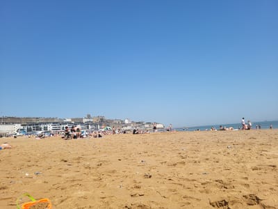 The image depicts a sandy beach on a sunny day in the city of Thanet, United Kingdom. The beach is populated with people, some of whom are sunbathing, walking, and relaxing. In the foreground is an orange and green plastic bucket. The sand is yellow-brown in color and shows footprints and variations in texture. In the background is a row of white buildings, including what appear to be apartment blocks, stretching across the horizon. The sky is a clear, light blue.