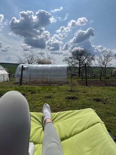 The image shows a sunny outdoor scene. The view is from a first-person perspective, looking down at a person sitting on a lime green cushion. The person is wearing light grey sweatpants and white socks. In the background, there is a green field with yellow flowers. There is a long greenhouse with a clear plastic covering running parallel to the camera. Beyond the greenhouse, there are bare trees and a field. The sky is blue with large, fluffy white clouds. The location is Lipnik, Bulgaria.