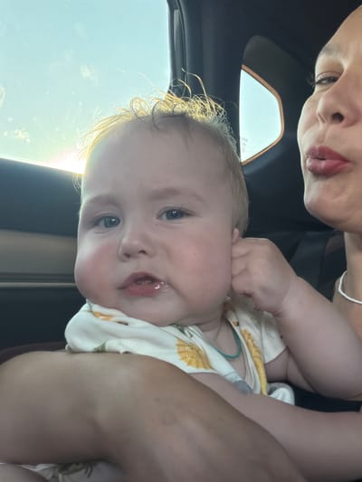 In this close-up, vertically oriented photograph, a baby is being held by an adult. The baby, with fair skin and blonde hair, looks directly at the camera with wide, blue eyes. Their mouth is slightly open, revealing a bit of tongue, and they have a small amount of drool on their chin. The baby is wearing a white onesie with a yellow floral pattern and a teal-colored string around their neck.
The adult's face is partially visible in the upper left corner of the frame. They have light skin and their lips are painted a deep red. Their expression is not fully discernible due to the angle and cropping.
The background suggests they are inside a vehicle. The upper portion of the image shows the interior of a car, possibly near the sunroof or ceiling. A sliver of bright sky with some clouds is visible through what appears to be a sunroof or window, indicating it is daytime and likely sunny. The lighting casts a warm glow on the baby's hair. The scene appears casual and intimate, likely a parent or caregiver capturing a moment with their child while on a journey. No visible text is present in the image.