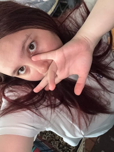 The image presents an overhead, close-up view of a person, likely female, lying down indoors in Lyman, Ukraine. The subject has fair skin and long, dark red hair fanned out around their head. They are wearing a white t-shirt, which appears somewhat rumpled, suggesting a relaxed posture on a bed or soft surface.

One of the person's hands is positioned over their face, with fingers splayed, partially obscuring the nose and the right eye (viewer's left). The visible eye and the partially covered eye are wide open, gazing directly at the camera, conveying an expression of surprise, curiosity, or playful candidness. Two small, silver ball piercings are visible on the person's left eyebrow.

The background consists of white fabric, likely a pillow or bedding, and a darker, patterned fabric visible on the far right and top, possibly a blanket, rug, or furniture, indicating a casual and intimate setting such as a bedroom or living area. No specific time of day, weather conditions, or visible text can be determined from the image.