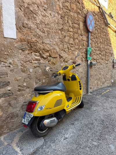 A yellow Vespa scooter is parked on a stone-paved surface against a stone wall in Deià, Spain. The scooter is positioned at an angle to the viewer with the rear of the vehicle closest to the wall. The rear license plate reads "0284 LYP." A circular blue and brown street sign on a metal pole, a small green and white rectangular sign, and electrical conduit are mounted on the stone wall. The image appears to be taken from above, looking down. A pair of shoes sits at the top of the image.