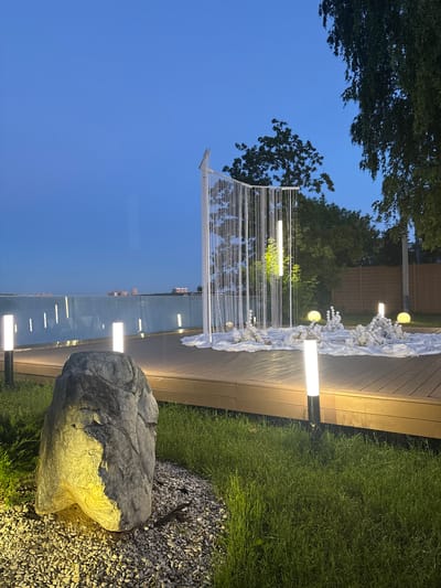 This image depicts an outdoor event space at dusk in Osipovo, Russia. The foreground features a large, illuminated boulder situated on a bed of small pebbles, with a neatly mowed lawn to its right. A wooden deck, bordered by a reflective glass barrier, extends into the mid-ground. Along the edge of the deck, several modern rectangular lamps illuminate the pathway.

On the deck, a decorative setup is arranged. This includes a white fabric draped across the surface, adorned with white floral arrangements and spherical lights. A tall, white structure with cascading white beaded strands stands as a central focal point. Two vertical, illuminated light fixtures are suspended from this structure. In the background, trees and a wooden fence are visible. Beyond the deck, a body of water is present, with distant buildings on the opposite shore under a deep blue sky. The overall scene suggests a tranquil, perhaps celebratory, ambiance illuminated by artificial lighting as daylight fades.