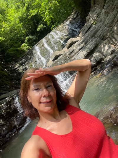 A woman is taking a selfie in a natural setting by a waterfall. She is wearing a red top and has her hand on her forehead, looking at the camera with a slight smile. The background shows a rocky cliff face with water cascading down it, surrounded by lush green foliage. The water appears clear and shallow in some areas. Sunlight is illuminating the scene, suggesting it is daytime. The overall impression is one of enjoying nature and relaxation in a beautiful outdoor environment.