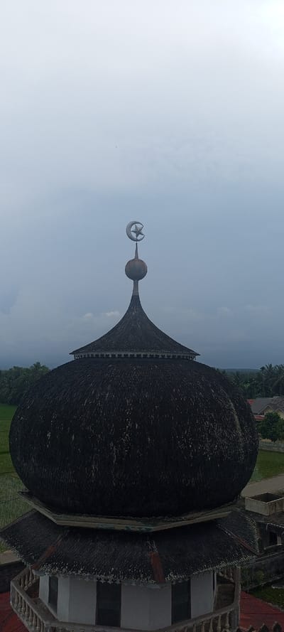 The image depicts the upper section of a mosque structure in Kumbang, Indonesia, captured from an elevated perspective under an overcast light-grey sky. The primary subject is a large, dark, bulbous dome, appearing multi-tiered, its surface textured with countless small, overlapping, dark elements, possibly shingles or tiles. A tapering spire rises from the apex of the dome, culminating in a metallic, spherical element surmounted by a prominent silver-colored finial shaped as a crescent moon enclosing a five-pointed star. Below the main dome, a secondary, smaller tiered roof, also dark and textured, features white decorative trim along its edge. This structure rests upon a lighter-colored section exhibiting windows and a balustrade-style railing. The background reveals a landscape of vibrant green rice paddies and dense tropical foliage, including numerous palm trees, extending towards the left. To the right, scattered residential buildings with orange or red roofs are visible amidst more trees. No individuals are present in the scene.