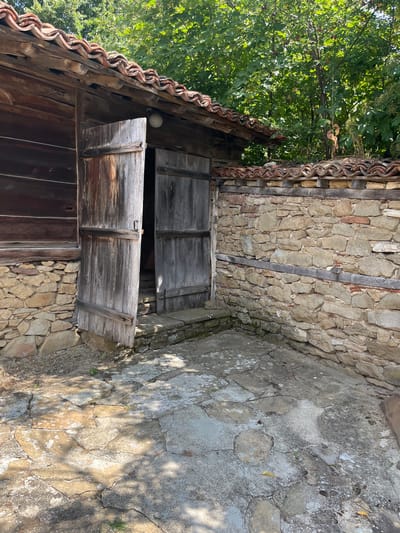 The image captures an exterior view of a traditional Bulgarian house in Zheravna, Bulgaria. The house features a wooden facade and a tiled roof. In the foreground, a stone wall encloses a courtyard paved with irregular stone slabs. The courtyard is partially shaded by trees. Two large wooden doors, one slightly ajar, are visible on the side of the house, leading into a dark interior. The overall scene depicts an old, rustic architectural style.