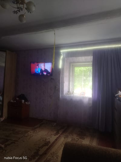 The image depicts an interior view of a room, likely a living room or bedroom. The room is sparsely furnished, with a television mounted on the wall displaying two men. A window with a sheer curtain and a thicker, purple curtain is visible, covered by a string of LED lights. A chest of drawers is positioned below the television, with items on top including a cup. The floor is covered by a patterned rug. A ceiling light fixture is visible in the upper left corner. The overall impression is of a simple, lived-in space, possibly in a rural setting given the greenery visible outside the window. There are no explicit indicators to confirm the location as Sestrynivka, Ukraine, however, if this is indeed the case, it would suggest a modest dwelling within that area.