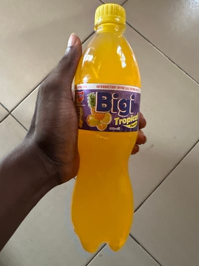 A dark-skinned left hand holds a transparent plastic bottle of "BiGi Tropical Flavor" beverage, filled with a yellow-orange liquid, in an indoor setting in Ketu Adieowe, Nigeria. The bottle features a yellow screw cap and a purple label with "BiGi" prominently displayed, along with "Tropical Flavor," "600mle," "INTRODUCTORY OFFER 20% EXTRA FREE," and graphics of a pineapple and citrus fruits. Condensation is visible on the bottle's exterior. The background consists of light-colored square tiles with dark grout lines.