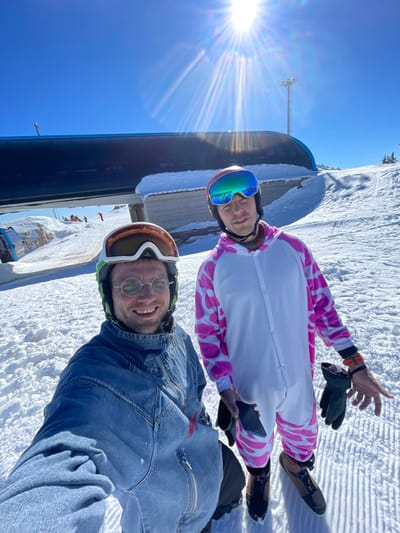 A selfie-style image captures two adult males standing on a snow-covered ski slope in Jahorina, Bosnia and Herzegovina, under a clear blue sky with a prominent sun causing lens flare. The male in the foreground, positioned on the left, wears a green ski helmet, round metal-framed sunglasses, and a blue denim-style zip-up jumpsuit or overall. He is smiling and appears to be holding the camera. The male on the right wears a red ski helmet with reflective blue-green goggles and a white full-body suit (onesie) patterned with pink splotches and front button closures. He holds a dark-colored smartphone in his left hand, and dark green gloves in his right hand, while wearing an orange digital watch on his left wrist. Both individuals are standing on groomed snow. In the background, a large, dark ski lift station or terminal is visible on the left, with distant figures on the slope and a tall metal utility pole to the right.
