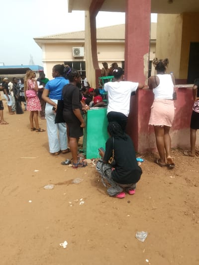The scene is an outdoor gathering on a dusty, reddish-brown ground adjacent to a building in Umuokpu, Nigeria. A woman in a black top, grey distressed pants, and pink sandals crouches in the foreground, facing away from the camera, holding a smartphone. A black shoulder bag with a gold chain strap hangs from her left shoulder. To her right, a woman in a white cropped tank top and a light pink mini-skirt, wearing brown flip-flops, stands with her back to the camera, leaning against a reddish-brown painted pillar of the building. Her hair is styled in two prominent buns. Immediately to her left, another woman in a white t-shirt and dark pants also leans against a similar reddish-brown pillar, her back to the camera. Further left, a woman in a black dress stands facing right, looking towards a green waist-height barrier. Beside her, a woman in a blue top and light-colored wide-leg pants stands, with a smartphone partially tucked into her left waistband. To her left, a woman in a red and white floral dress with a pink shoulder bag stands, looking towards the crowd. A man in a green t-shirt is partially obscured behind her.
Numerous other individuals, predominantly women, are visible in the midground and background. Some are seated on green chairs behind the green barrier, while others stand in groups. The background features a light-colored building with a beige facade and reddish-brown painted lower sections and pillars. An air conditioning unit is mounted on the building's exterior. In the far left background, a large vehicle, possibly a bus, is partially visible, near a blue, elevated industrial structure. The ground is characterized by scattered litter, including plastic wrappers and torn paper.
