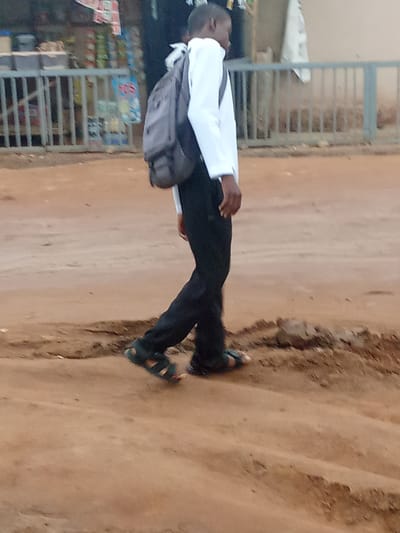 A person is walking on a dirt road in Kagini, Nigeria. The person is wearing a white long-sleeved shirt, black pants, and sandals. They are also carrying a gray backpack. In the background, there is a building with shelves and various items on display. There is also a metal fence. The ground is dry and dusty, with some uneven patches. The overall scene depicts an outdoor environment in a Nigerian urban or semi-urban setting.