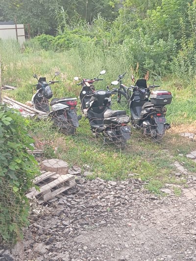 The image depicts an outdoor scene with several parked electric scooters in a grassy area. In the foreground, a chain-link fence partially obscures the view, with green foliage on the left and gravelly ground with scattered stones and debris. Behind the fence, the electric scooters are arranged in a cluster. There are at least four scooters visible, some black and some white and black, each with a storage box or seat on the rear. The scooters are parked on uneven, grassy terrain. In the background, dense green foliage and trees form a natural backdrop. A white fence structure is partially visible on the far left. The scene suggests a storage or parking area for these vehicles, possibly in a semi-rural or less-developed urban setting within Novopavlovka, Kyrgyzstan. No people are visible in the frame.