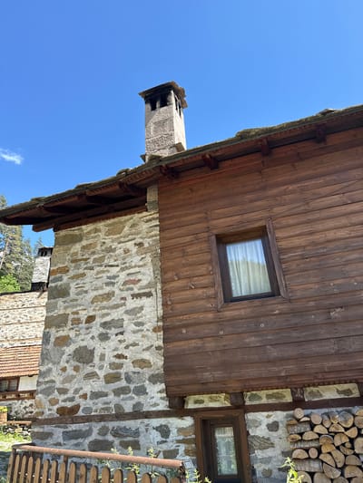 The image shows a traditional Bulgarian house in the village of Shiroka Laka. The house has a stone foundation with wooden upper walls. The roof is made of stone slabs. There is a chimney made of stone on the roof. A window with white curtains is visible on the wooden wall. In front of the house, there is a pile of firewood. The sky is clear and blue. The overall impression is of a rustic and charming rural dwelling.