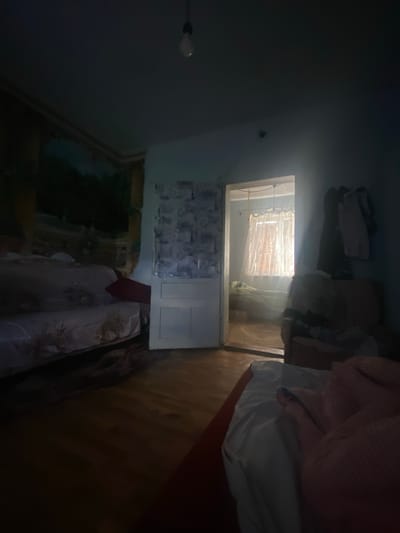The image depicts a dimly lit bedroom in a dwelling located in Borshchiv, Ukraine. The room appears to be in a state of disarray. In the foreground, a bed is visible with rumpled white and pink bedding. To the left, another bed or sofa is partially covered with a patterned fabric, and a red pillow is propped up against it. A large tapestry with a landscape scene hangs on the wall to the left. A doorway in the center of the image leads to an adjacent room where a window with sheer curtains is visible, allowing light to filter through. Clothes are hanging on the right side of the doorway. The floor is made of wooden planks. A bare light bulb hangs from the ceiling. The overall atmosphere is dark and somewhat neglected.